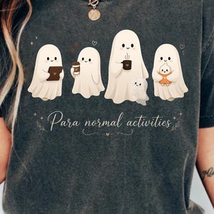 Peut inclure: Un t-shirt gris anthracite orné d'un motif représentant quatre fantômes de dessin animé effectuant des tâches quotidiennes, tenant une tablette, une tasse de café et un plus petit fantôme. Le texte "Para normal activities" est inscrit en dessous.