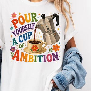 Peut inclure: Un t-shirt blanc avec un motif coloré représentant une cafetière italienne argentée versant du café dans une tasse assortie. Le texte "POUR YOURSELF A CUP OF AMBITION" est écrit en lettres multicolores, entouré de petites fleurs et d'étoiles.