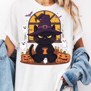 May include: A white t-shirt featuring a graphic of a black cat wearing a purple witch hat, holding a mug. The cat is surrounded by orange jack-o'-lanterns, lit candles, and small white ghosts against a backdrop of a window with a yellow sky.