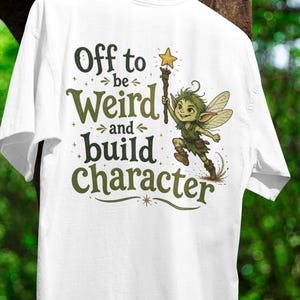Peut inclure: Un t-shirt blanc à manches courtes avec une illustration d'une petite fée verte tenant une baguette surmontée d'une étoile. Le texte "Off to be Weird and build Character" est écrit en vert foncé, entouré de petites étoiles.