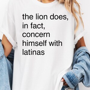 Peut inclure: Un t-shirt blanc à manches courtes avec un texte noir imprimé sur le devant qui dit, the lion does, in fact, concern himself with latinas. Le t-shirt a une encolure ronde et une coupe décontractée, associé à une veste en jean bleu clair.