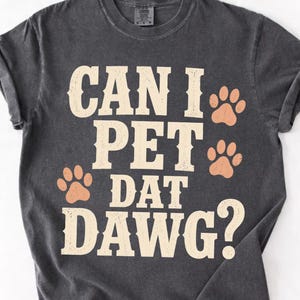 Peut inclure: Un t-shirt gris anthracite à manches courtes avec le texte crème "CAN I PET DAT DAWG?" imprimé sur le devant. Le motif comprend quatre empreintes de pattes orange clair entourant le texte, écrit dans une police serif grasse et usée.