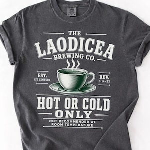 May include: A charcoal gray short-sleeve t-shirt featuring a white graphic of a coffee cup on a saucer. The text reads: THE LAODICEA BREWING CO. EST. 1ST CENTURY REV. 3:14-22 HOT OR COLD ONLY NOT RECOMMENDED AT ROOM TEMPERATURE.
