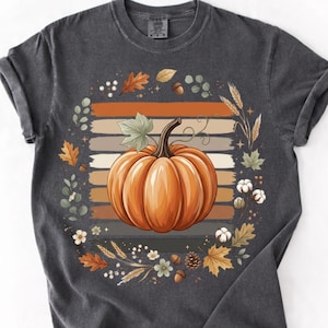 Peut inclure: Un t-shirt à manches courtes gris anthracite avec un motif central représentant une grosse citrouille orange. La citrouille est entourée d'une couronne de feuilles d'automne, d'épis de blé, de glands et de petites fleurs blanches.