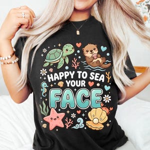 Peut inclure: Un t-shirt noir à manches courtes avec un motif coloré représentant une tortue de mer, une loutre, une étoile de mer et un coquillage avec une perle. Le texte "HAPPY TO SEA YOUR FACE" est écrit en grandes lettres bleu clair avec des cœurs.