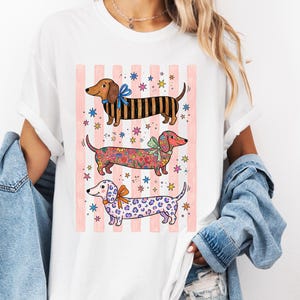Peut inclure: Un t-shirt blanc à manches courtes avec une impression graphique représentant trois chiens teckels. Le chien du haut a des rayures noires, celui du milieu un motif floral et celui du bas un imprimé léopard violet. Chaque chien porte un nœud sur un fond rayé rose.