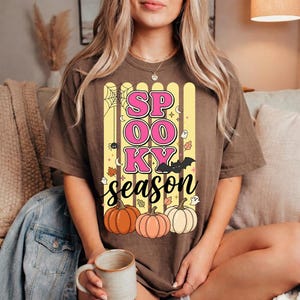 Peut inclure: Un t-shirt marron avec une impression graphique portant le texte "SPOOKY season" en rose et noir. Le motif comprend des rayures verticales jaunes, une toile d'araignée, une araignée, une chauve-souris, des petits fantômes, des feuilles d'automne et trois citrouilles orange et blanches.