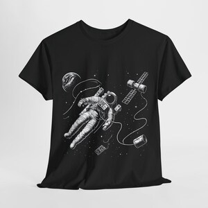 Könnte beinhalten: Schwarzes T-Shirt mit einem weißen Grafikdruck eines Astronauten, der im Weltraum schwebt, mit einer Raumstation und Sternen im Hintergrund.