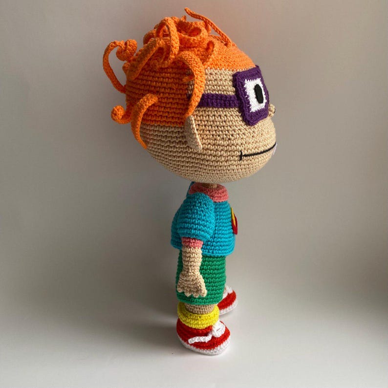 May include: A crocheted plush toy of a cartoon character with orange hair, purple glasses, a blue shirt, green shorts, and red shoes.