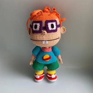Könnte beinhalten: Häkelpuppe Amigurumi eines Comic-Charakters mit orangefarbenem Haar, lila Brille und einem blauen Shirt mit einem roten Planetenmuster. Die Puppe trägt rote und gelbe Schuhe.