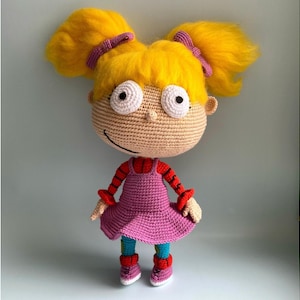 May include: Crocheted doll with yellow hair, pink dress, and red and white striped sleeves. The doll has large white eyes and a pink nose.