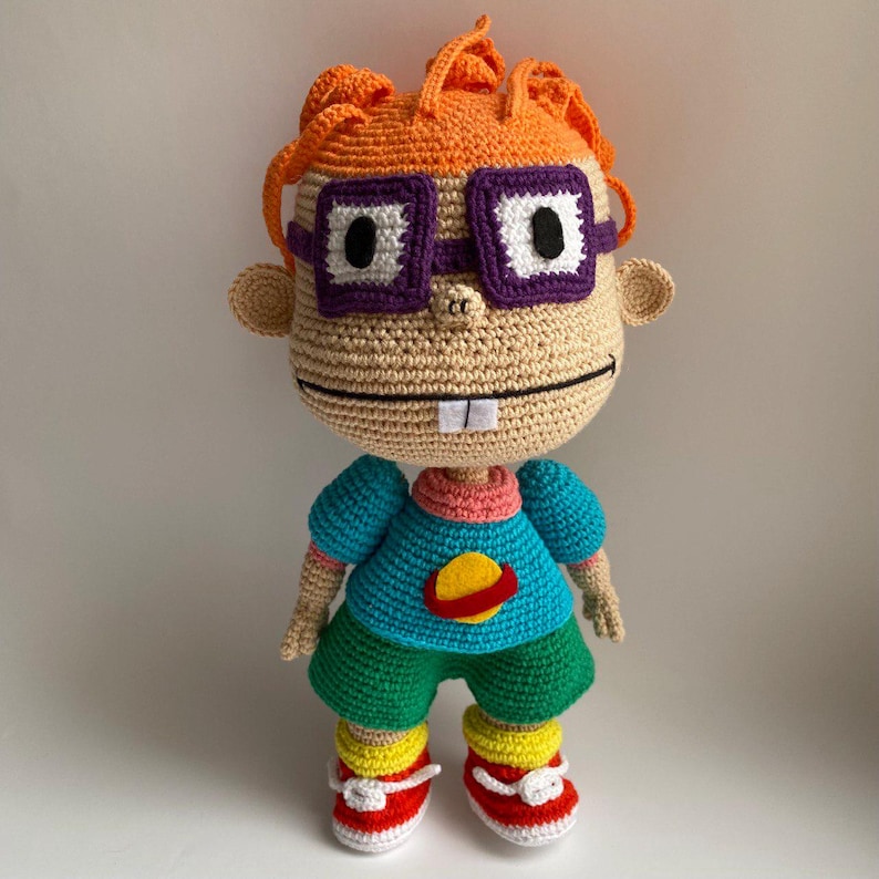 May include: A crocheted plush toy of a cartoon character with orange hair, purple glasses, and a blue shirt with a yellow planet design. The toy is wearing green trousers and red and yellow shoes.