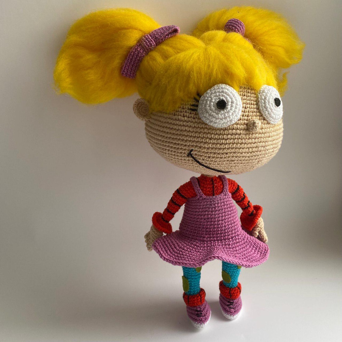 May include: A crocheted doll with yellow hair, large white eyes, and a pink dress. The doll is wearing red and black striped sleeves and blue and green patterned leggings.