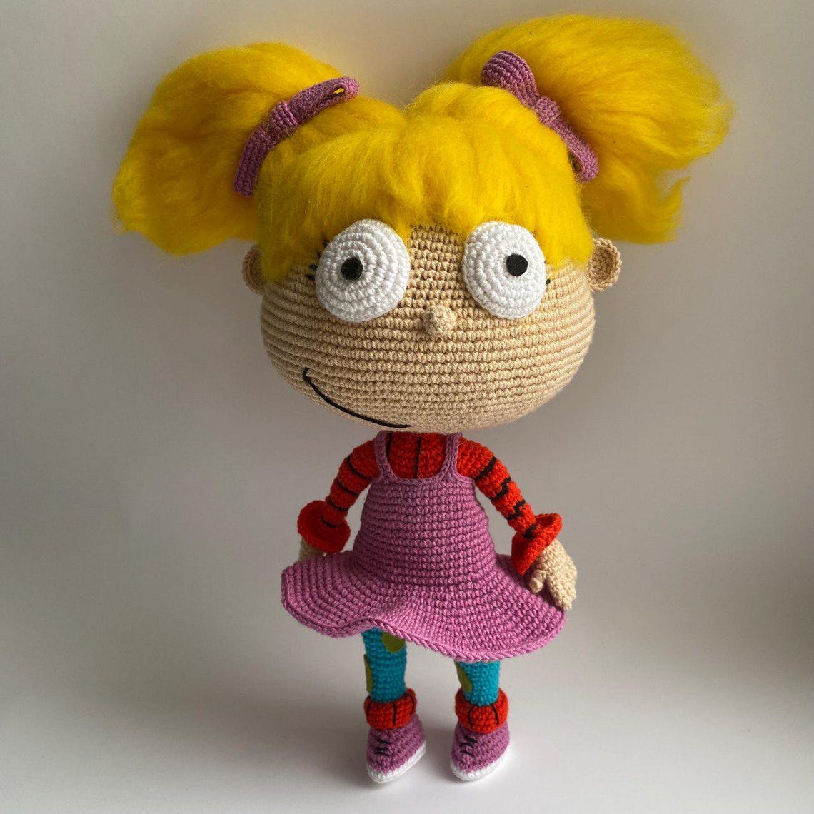 May include: A crocheted doll with yellow hair, large white eyes, and a pink dress. The doll is wearing red and white striped sleeves and blue and white striped leggings.