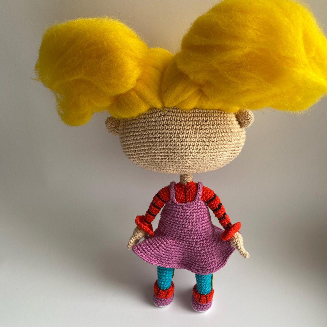 May include: A crocheted doll with yellow yarn hair, a pink dress, and red and black striped sleeves. The doll is standing on a white surface.