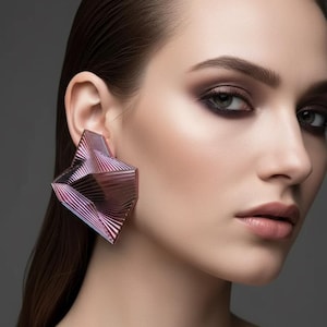 May include: Geometric, angular earrings in shades of pink and purple. The earrings have a layered, three-dimensional design. The image is a close-up of a person's face, with the earrings as the focal point. The background is a solid gray.