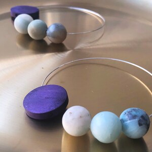 May include: Two pairs of hoop earrings with a silver wire and a series of round beads. One pair has a purple wooden bead and three light blue beads. The other pair has a dark purple wooden bead and three light blue beads.