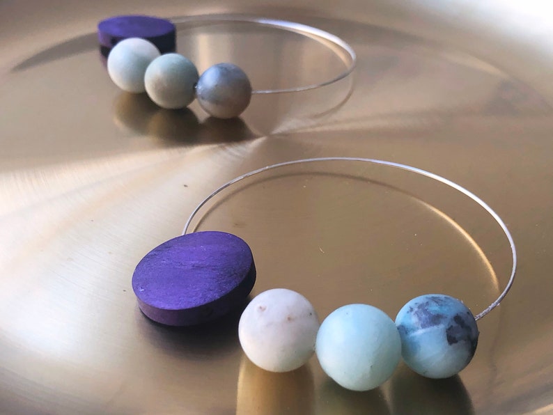 May include: Two pairs of hoop earrings with a silver wire and a series of round beads. One pair has a purple wooden bead and three light blue beads. The other pair has a dark purple wooden bead and three light blue beads.