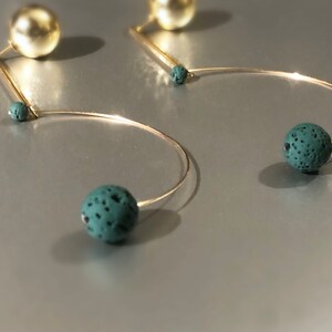 May include: Gold-toned earrings with a unique design. Each earring features a large, spherical gold stud, a thin gold bar, and a curved wire ending in a speckled, teal-colored sphere. The earrings have a modern, minimalist aesthetic.
