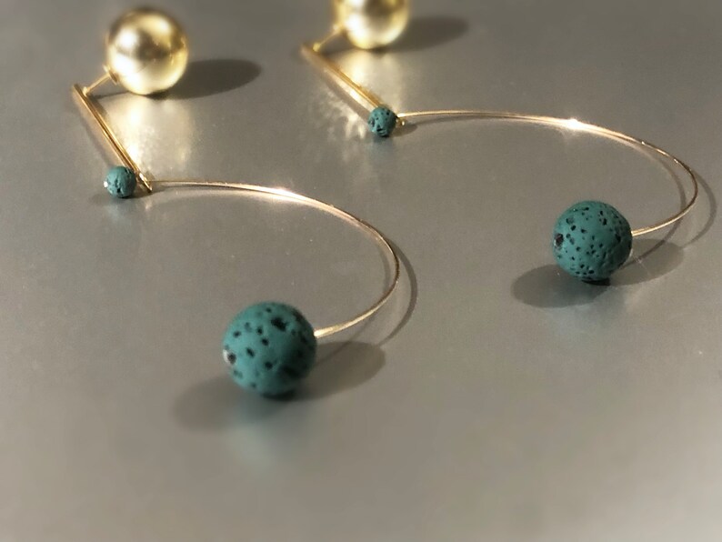 May include: Gold-toned earrings with a unique design. Each earring features a large, spherical gold stud, a thin gold bar, and a curved wire ending in a speckled, teal-colored sphere. The earrings have a modern, minimalist aesthetic.