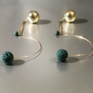 May include: A pair of unique earrings featuring a gold-tone wire design. Each earring has a large, gold-colored sphere at the top, a small teal sphere, and a larger teal sphere at the bottom. The earrings have a modern, abstract style.