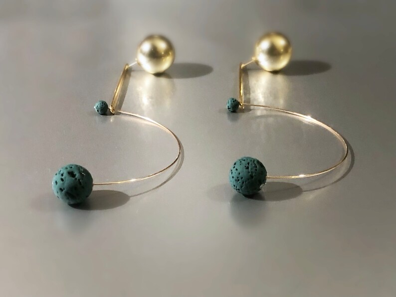 May include: A pair of unique earrings featuring a gold-tone wire design. Each earring has a large, gold-colored sphere at the top, a small teal sphere, and a larger teal sphere at the bottom. The earrings have a modern, abstract style.