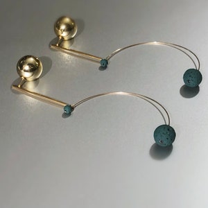 May include: A pair of gold-toned earrings. Each earring features a large, round gold ball at the top, a straight gold bar, a small teal bead, a curved silver wire, and a textured teal ball at the end. The earrings are displayed on a silver surface.