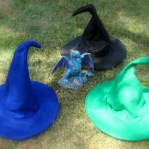 Pu&ograve; includere: Tre cappelli da strega di diversi colori: blu, nero e verde. I cappelli sono sdraiati su un letto di erba verde. Una piccola statuetta di drago blu si trova al centro dell'immagine.
