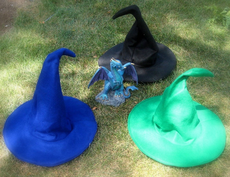 Pu&ograve; includere: Tre cappelli da strega di diversi colori: blu, nero e verde. I cappelli sono sdraiati su un letto di erba verde. Una piccola statuetta di drago blu si trova al centro dell'immagine.