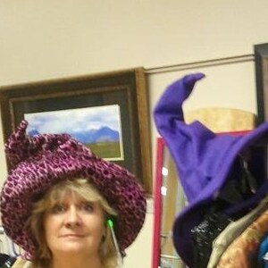 Pu&ograve; includere: Una donna indossa un cappello rosa e viola con un'ampia tesa. Il cappello ha una punta appuntita ed &egrave; decorato con una piccola piuma verde. La donna sorride e guarda la telecamera.