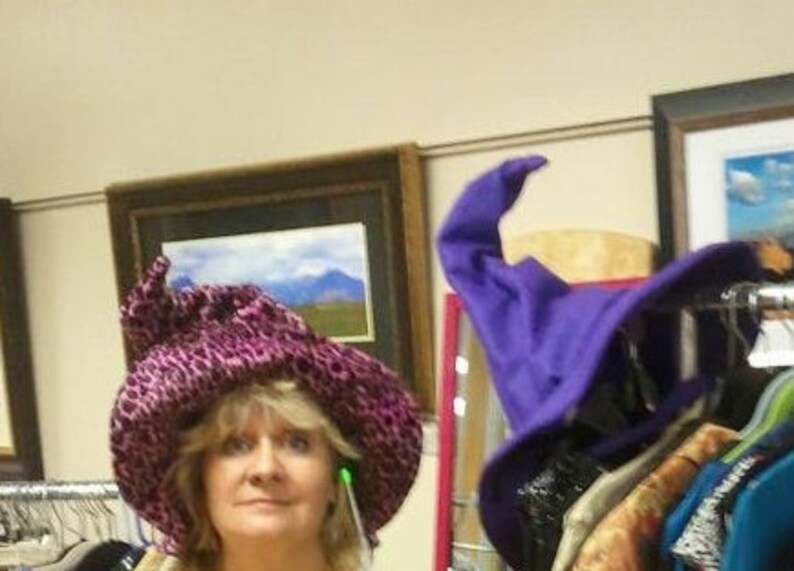 Pu&ograve; includere: Una donna indossa un cappello rosa e viola con un'ampia tesa. Il cappello ha una punta appuntita ed &egrave; decorato con una piccola piuma verde. La donna sorride e guarda la telecamera.