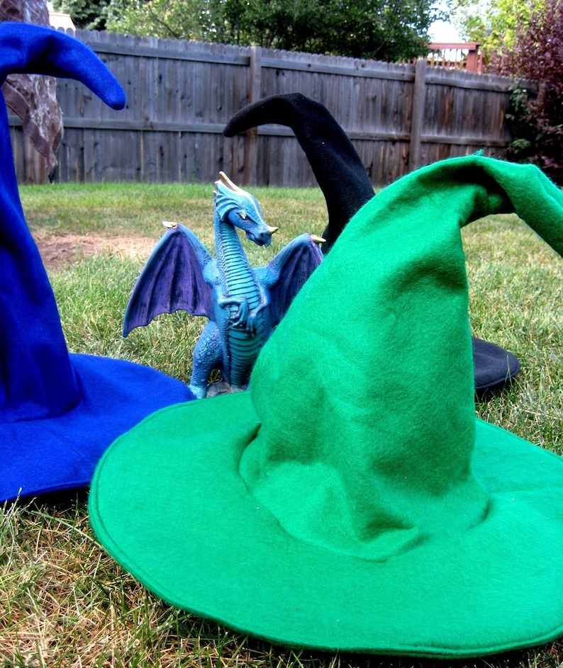 Pu&ograve; includere: Tre cappelli da strega, uno blu, uno nero e uno verde, sono disposti su una superficie erbosa. Una piccola statuetta di drago viola e blu si trova al centro dell'immagine.