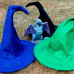Pu&ograve; includere: Tre cappelli da strega in blu, verde e nero. I cappelli sono fatti di feltro e hanno un'ampia tesa. Una statuetta di drago blu si trova al centro dell'immagine.