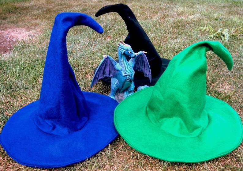 Pu&ograve; includere: Tre cappelli da strega in blu, verde e nero. I cappelli sono fatti di feltro e hanno un'ampia tesa. Una statuetta di drago blu si trova al centro dell'immagine.