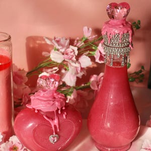 May include: Two pink decorative bottles with heart-shaped accents. One bottle is heart-shaped with a crystal heart and a dripping pink wax design. The other is a champagne bottle shape with a crystal heart and a rhinestone collar.