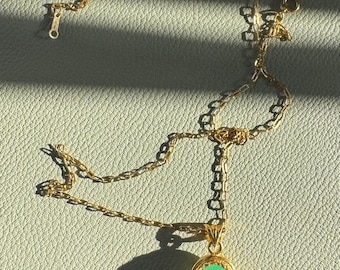 Op de afbeelding: Een goudkleurige ketting met een hanger met een lichtgroene ovale steen. De ketting is delicaat en de hanger is gezet in een gouden bezel. De ketting wordt gepresenteerd op een getextureerd, lichtgroen oppervlak.