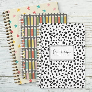 May include: Three spiral bound notebooks with different designs. The first notebook has a white background with colorful stars. The second notebook has a gray background with colorful pencils. The third notebook has a white background with black dalmatian spots. The third notebook has a label that says "Mrs. Thompson, Washington Elementary School".