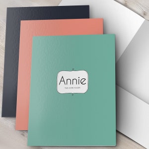 May include: A teal take-home folder with the name "Annie" written in a white oval with a black outline. The folder is stacked on top of a coral folder and a black folder.