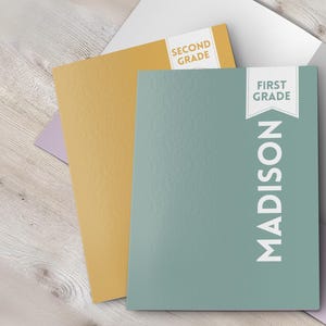 May include: A green folder with the name "MADISON" printed vertically in white. The folder has a white tab with the text "FIRST GRADE" printed on it. The folder is open to reveal a white interior and a yellow folder with the text "SECOND GRADE" printed on it.