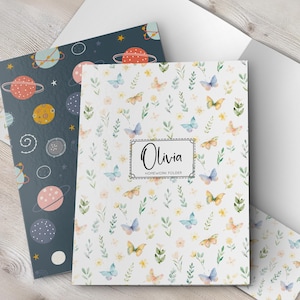 May include: Two folders, one with a navy blue space theme and the other with a white butterfly and floral pattern. The butterfly folder has the name "Olivia" and "Homework Folder" printed on it. The folders are on a light wood surface.