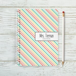 May include: A spiral bound notebook with a colourful striped cover. The cover has diagonal stripes in shades of pink, yellow, and green. The notebook has a label that says "Mrs. Tiernan, School Principal".