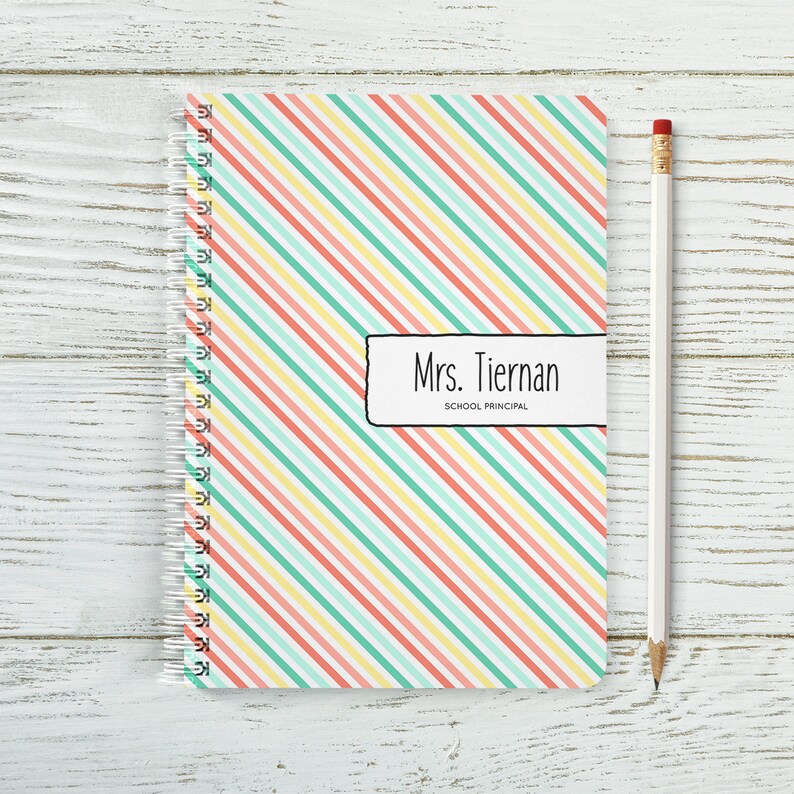 May include: A spiral bound notebook with a colourful striped cover. The cover has diagonal stripes in shades of pink, yellow, and green. The notebook has a label that says "Mrs. Tiernan, School Principal".