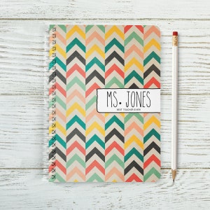 May include: A spiral bound notebook with a colourful chevron pattern in shades of pink, yellow, green, blue, and black. The notebook has a white label with the text "MS. JONES BEST TEACHER EVER!"
