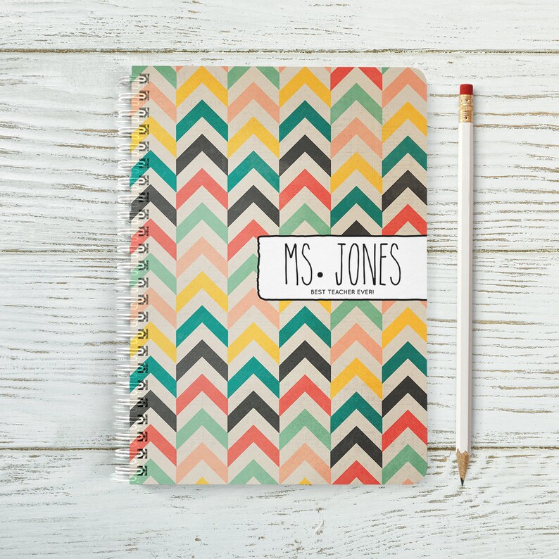 May include: A spiral bound notebook with a colourful chevron pattern in shades of pink, yellow, green, blue, and black. The notebook has a white label with the text "MS. JONES BEST TEACHER EVER!"
