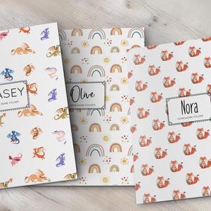 K&ouml;nnte beinhalten: Drei rechteckige Mappen mit wei&szlig;em Hintergrund und verschiedenen Mustern. Die erste zeigt bunte Drachen und den Text CASEY TAKE HOME FOLDER. Die zweite hat Regenb&ouml;gen und Sonnen mit dem Text Olive HOMEWORK FOLDER. Die dritte zeigt F&uuml;chse mit dem Text Nora HOMEWORK FOLDER.