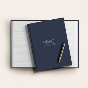 Może przedstawiać: Niebieski notes w kolorze granatowym z napisem "CHARLIE" i tekstem "FOR ADVENTURES, BIG & SMALL" na okładce. Na notesie leży złoty długopis.