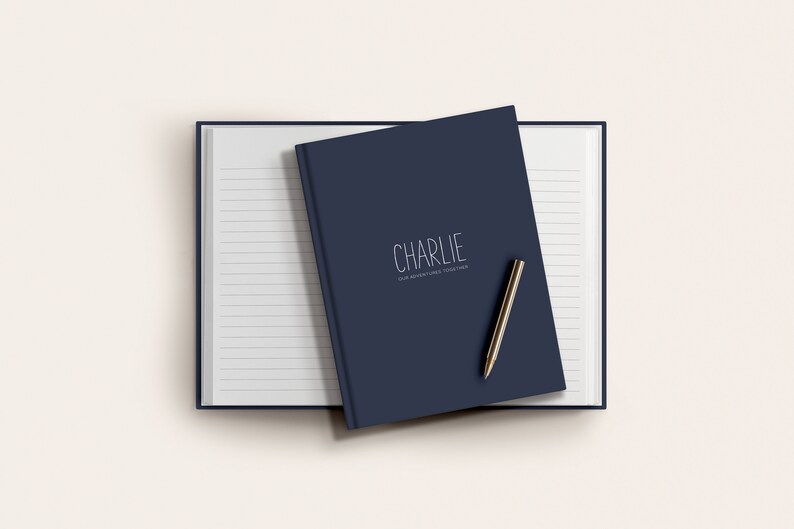 Może przedstawiać: Niebieski notes w kolorze granatowym z napisem "CHARLIE" i tekstem "FOR ADVENTURES, BIG & SMALL" na okładce. Na notesie leży złoty długopis.