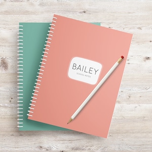 May include: A pink and green spiral notebook with the name "BAILEY" and the words "SCHOOL NOTES" printed on the front. A white pencil is lying on top of the notebook.
