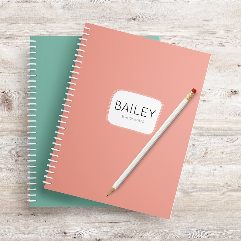 May include: A pink and green spiral notebook with the name "BAILEY" and the words "SCHOOL NOTES" printed on the front. A white pencil is lying on top of the notebook.