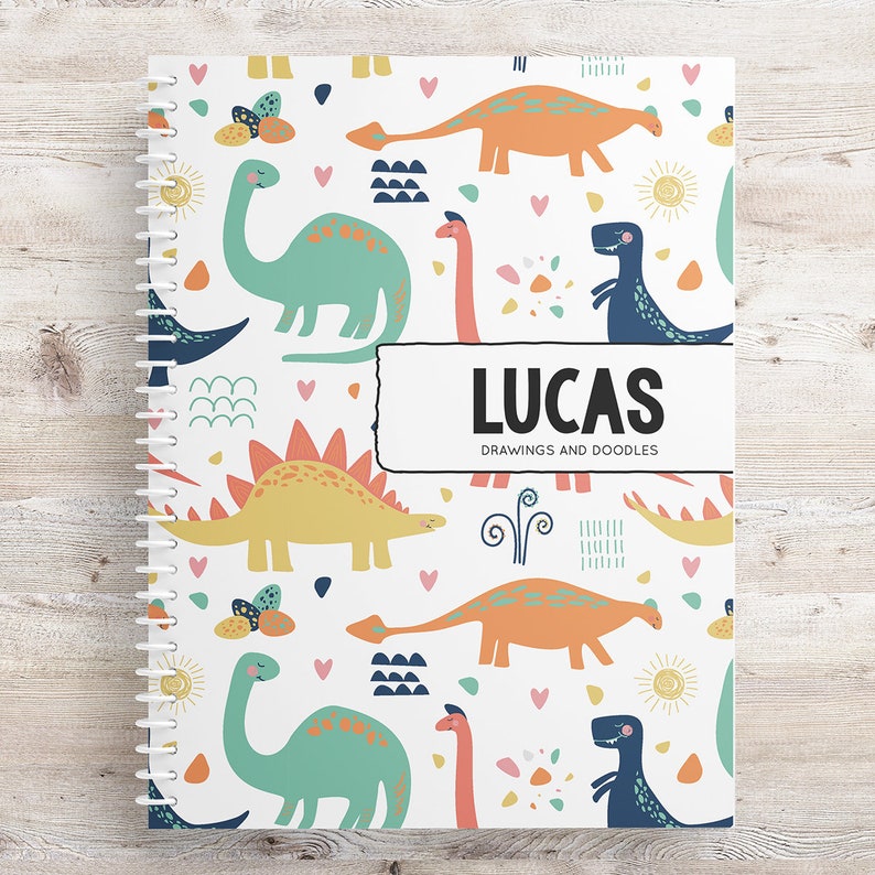 May include: A white spiral bound notebook with a colorful dinosaur pattern. The cover features a blue, green, orange, and yellow dinosaur design with hearts, suns, and other geometric shapes. The text "LUCAS DRAWINGS AND DOODLES" is printed on the cover.